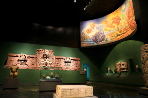 Ein Museumsraum mit verschiedenen Artefakten, darunter Skulpturen, Tischobjekte, eine Texttafel, eine Wandmalerei und Deckenbeleuchtung im Anthropologischen Museum von Mexiko-Stadt.