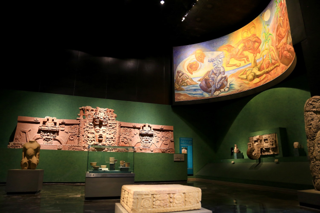 Ein Museumsraum mit verschiedenen Artefakten, darunter Skulpturen, Tischobjekte, eine Texttafel, eine Wandmalerei und Deckenbeleuchtung im Anthropologischen Museum von Mexiko-Stadt.