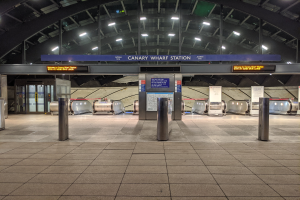Eine U-Bahn-Station mit Metalldetektoren auf dem Boden, Pollern, Informationsschildern mit der Aufschrift "Canary Wharf Station", Türen, Wänden und Deckenlampen.