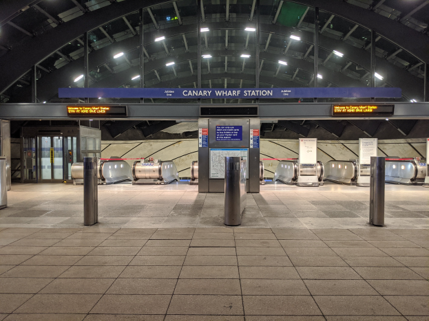 Eine U-Bahn-Station mit Metalldetektoren auf dem Boden, Pollern, Informationsschildern mit der Aufschrift "Canary Wharf Station", Türen, Wänden und Deckenlampen.