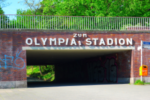 Der Eingang zum Olympiastadion in Berlin, Deutschland, mit einer Brücke mit Text, einem Metallzaun, einer Schautafel, einer Box, Pflanzen, Gras, einer Gruppe von Bäumen und einem bewölkten Himmel.