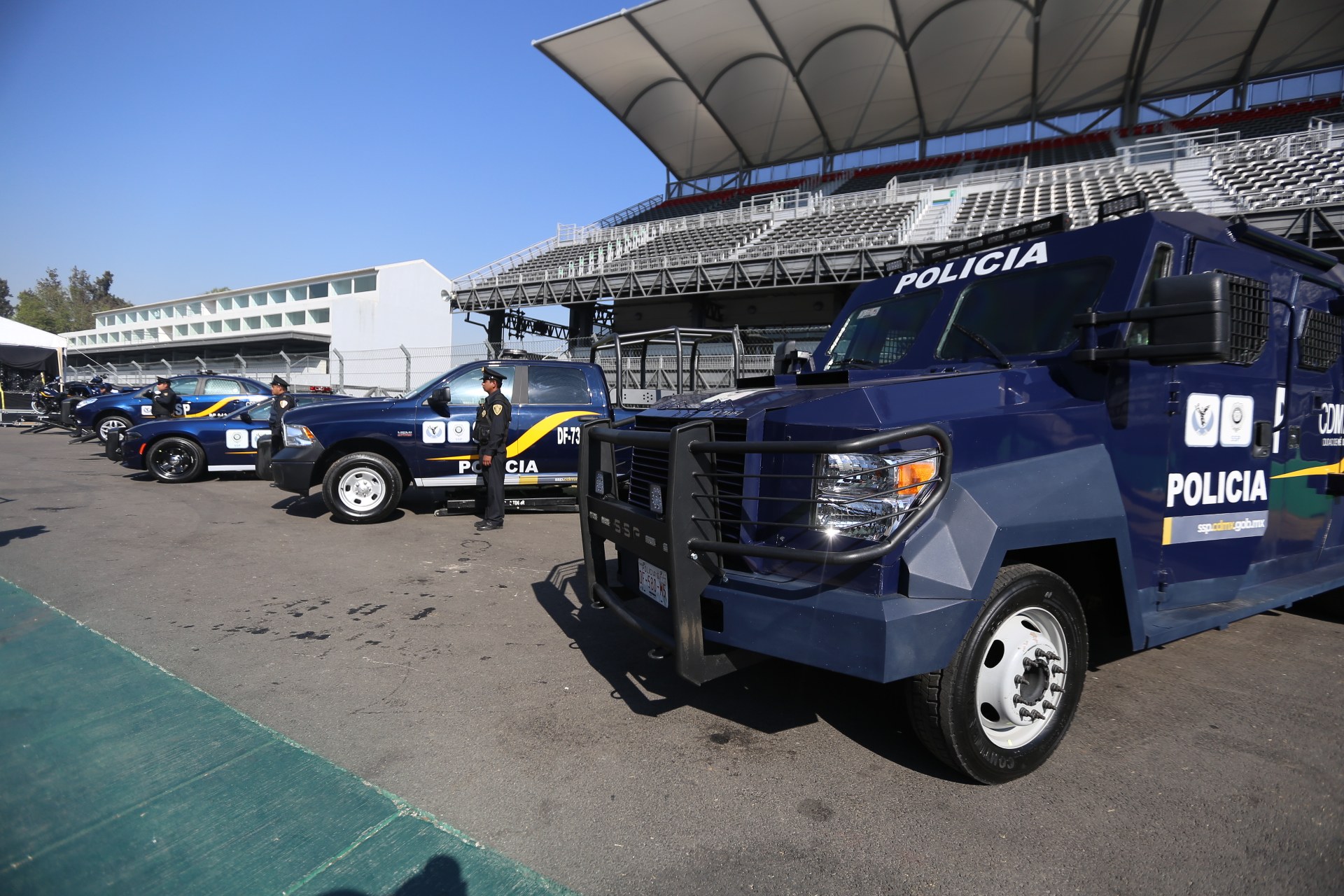 Eine Reihe von Polizeiwagen vor einem Stadion geparkt, mit Menschen im Vordergrund auf der Straße stehend, Gebäuden und Bäumen im Hintergrund und einem klaren blauen Himmel.