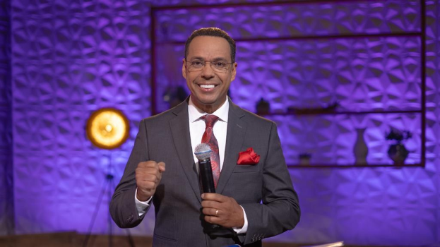 Stephen Colbert, Moderator von *The Daily Show with Trevor Noah*, steht auf einer Bühne und hält ein Mikrofon, trägt einen Anzug, Krawatte und Brille, mit einer Lampe und Ständer hinter ihm.