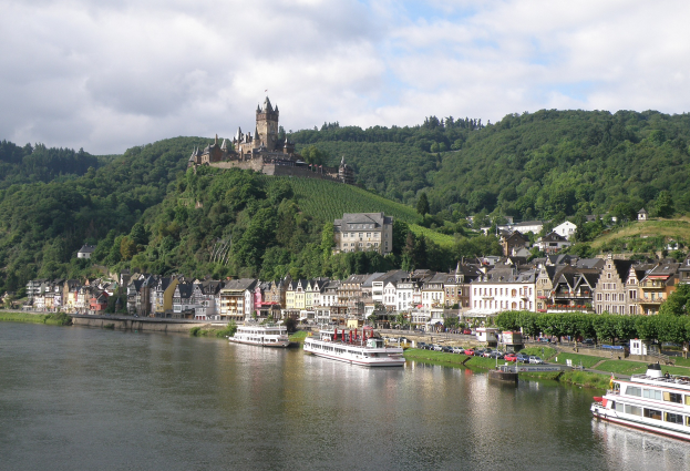 Ein malerischer Blick auf den Rhein in Deutschland mit einer Burg auf einem Hügel, Booten auf dem Fluss, Fahrzeugen auf einer nähergelegenen Straße und einem bewölktem Himmel.