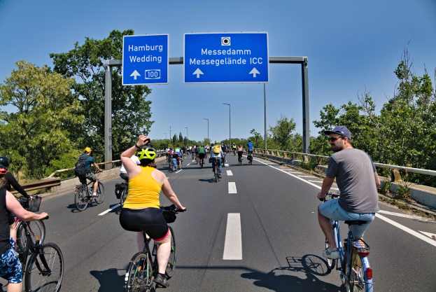 Gruppe von Radfahrern mit Helmen, die eine Straße mit Geländer und Bäumen auf beiden Seiten sowie Laternen im Hintergrund und einem klaren blauen Himmel hinunterfahren, mit einem Schild oben, das eine Radtour in Hamburg anzeigt.
