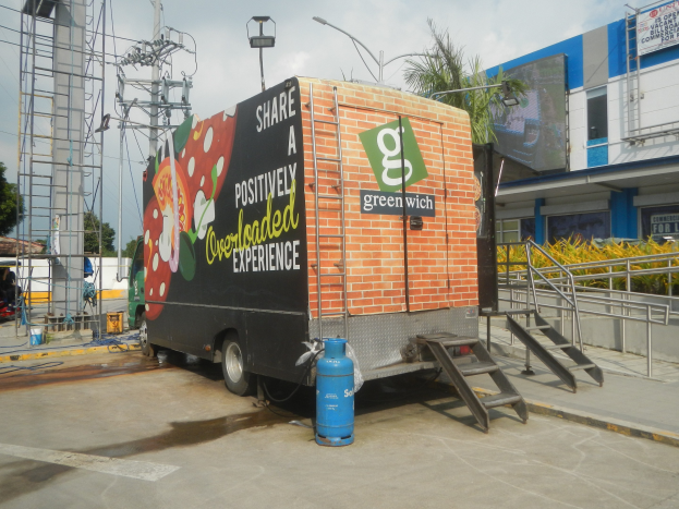 Ein Lkw ist vor einem Gebäude mit einer Tafel geparkt, auf der "Teile eine positiv überladene Erfahrung" steht, mit Stufen, die hinaufführen, einem Zylinder auf dem Boden, Geländern, Strommasten mit Drähten, Laternen, Bäumen und einem bewölkten Himmel im Hintergrund.