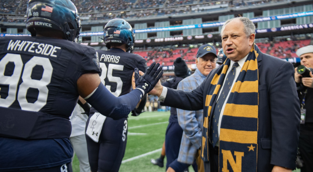 Ein Mann im Anzug und Krawatte gibt einem Fußballspieler die Hand auf einem Feld, umgeben von einer Gruppe von Menschen, mit einer Kamera Person rechts und einem Stadion voller Zuschauer im Hintergrund, das Schilder trägt, die "Notre Dame Football Coach Mike McCarthy und die Fighting Irish" anzeigen.