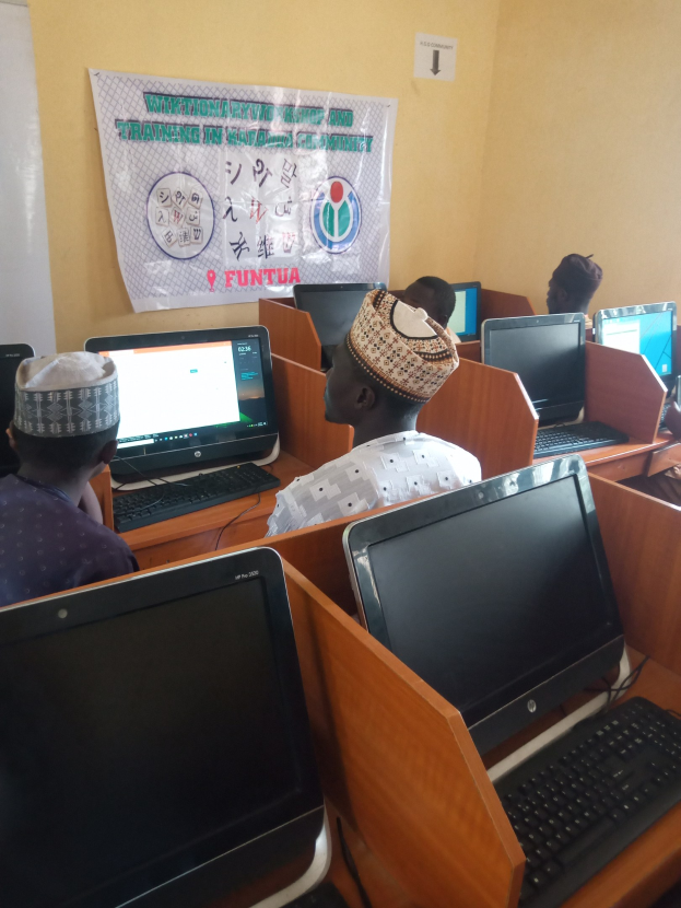 Eine Gruppe von Menschen sitzt an Schreibtischen mit Computerbildschirmen, Tastaturen und Mäusen und nimmt an einem Workshop zur Computertechnologie teil, wobei ein Plakat im Hintergrund zu sehen ist.