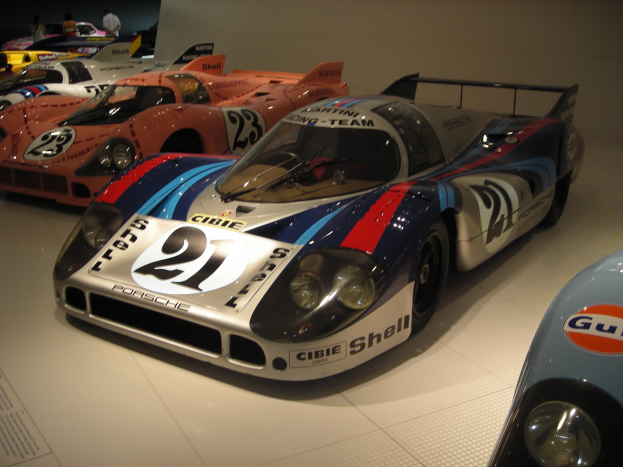 Ein Porsche 917K Rennwagen auf einer Museumsausstellung umgeben von anderen Sportwagen, mit Menschen in der Nähe und einer Wand im Hintergrund; ein Blatt Papier mit Text ist links sichtbar.