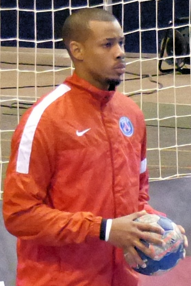 Ein Mann in einem blauen Hemd und Shorts steht vor einem Fußballtor und hält einen Fußball mit entschlossener Miene.