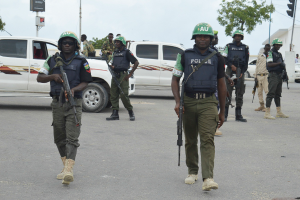 Eine Gruppe von Polizisten in Helmen und mit Waffen in der Hand geht eine Straße entlang mit Fahrzeugen, Laternen, Ampeln, Texttafeln, Bäumen, Gebäuden und einem bewölkten Himmel im Hintergrund.