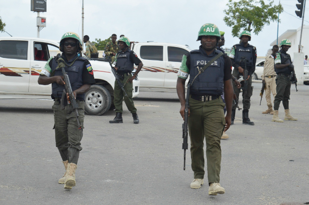 Eine Gruppe von Polizisten in Helmen und mit Waffen in der Hand geht eine Straße entlang mit Fahrzeugen, Laternen, Ampeln, Texttafeln, Bäumen, Gebäuden und einem bewölkten Himmel im Hintergrund.
