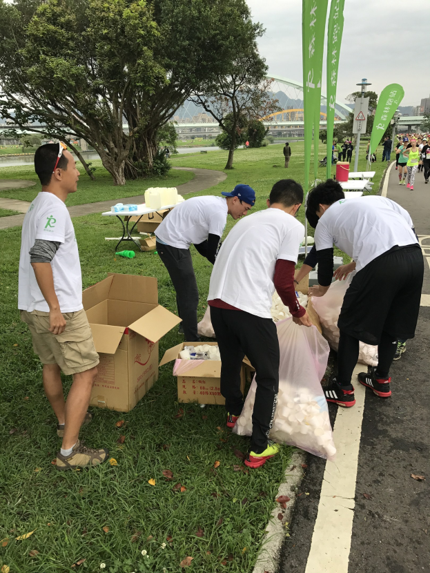 Group of people on a roadside with scattered cardboard boxes and bottles, participating in a charity run under clear blue sky with trees and buildings in the background.