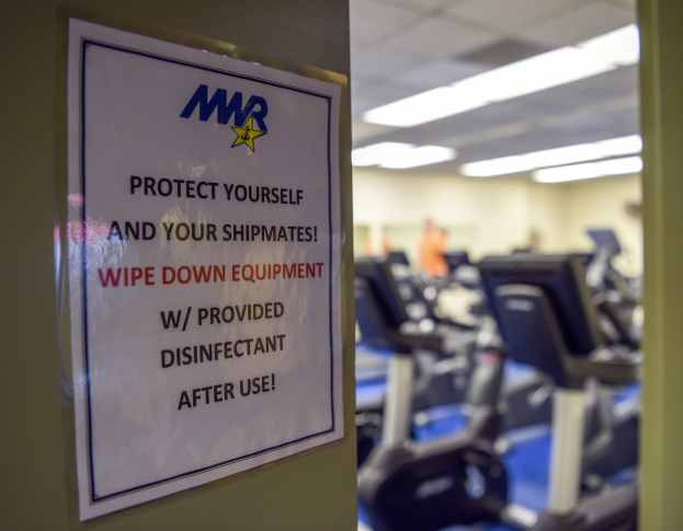 Ein Türschild mit der Aufschrift "Schützen Sie sich und Ihre Schiffskameraden" mit Laufbändern und Deckenleuchten im Hintergrund.