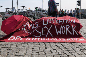 Gruppe von Menschen, die auf einer Stadtstraße protestieren und Regenschirme sowie ein rotes Banner mit der Aufschrift "Befreiung ohne Entkriminalisierung der Sexarbeit" halten, auf dem Bürgersteig.
