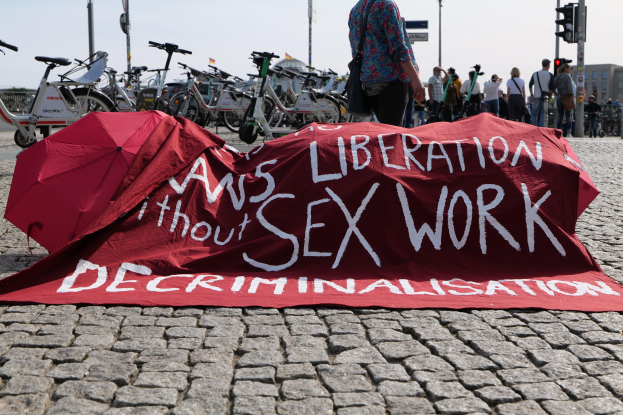 Gruppe von Menschen, die auf einer Stadtstraße protestieren und Regenschirme sowie ein rotes Banner mit der Aufschrift "Befreiung ohne Entkriminalisierung der Sexarbeit" halten, auf dem Bürgersteig.