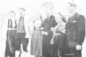 Eine Gruppe von Menschen in den 1930er Jahren, die in historischer Kleidung posieren, stehen zusammen im Freien mit Bäumen im Hintergrund, lächeln in die Kamera, mit dem Text 'Bayerische Familie in den 1930er Jahren' unten auf dem Bild.