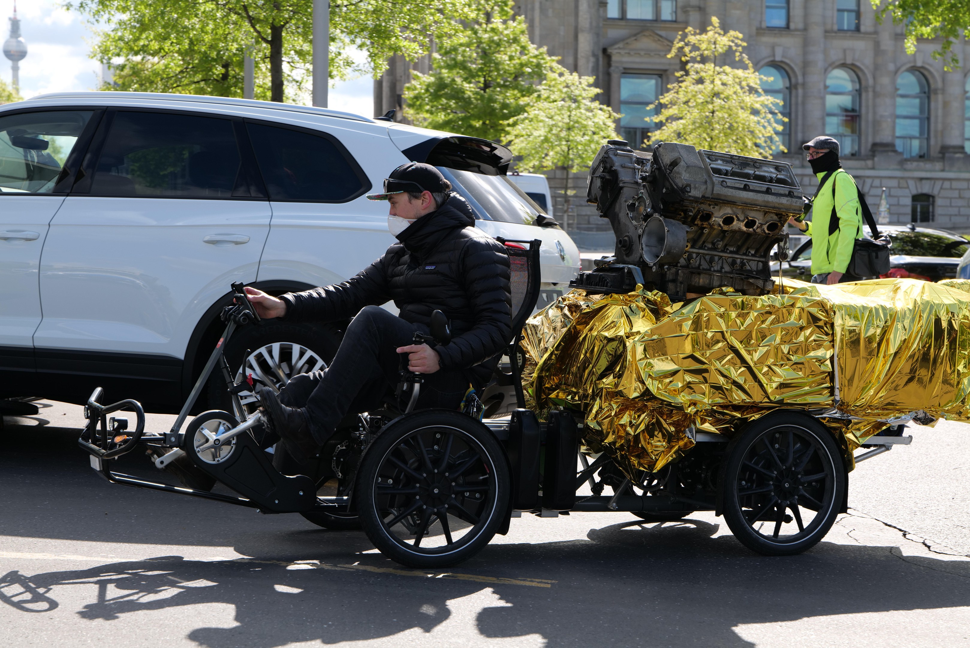 Ein Mann im Rollstuhl mit einem großen Motor auf dem Rücken, umgeben von Fahrzeugen auf einer Straße mit Bäumen, Gebäuden, Polen und einem klaren blauen Himmel im Hintergrund; er trägt eine schwarze Jacke, eine Mütze und hält ein Objekt.