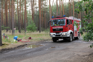 Ein rotes Feuerwehrauto fährt auf einer Schotterstraße durch den Wald, umgeben von Gras, einem Rohr und anderen Gegenständen auf der linken Seite der Straße, sowie weiteren Bäumen und einem klaren blauen Himmel im Hintergrund.