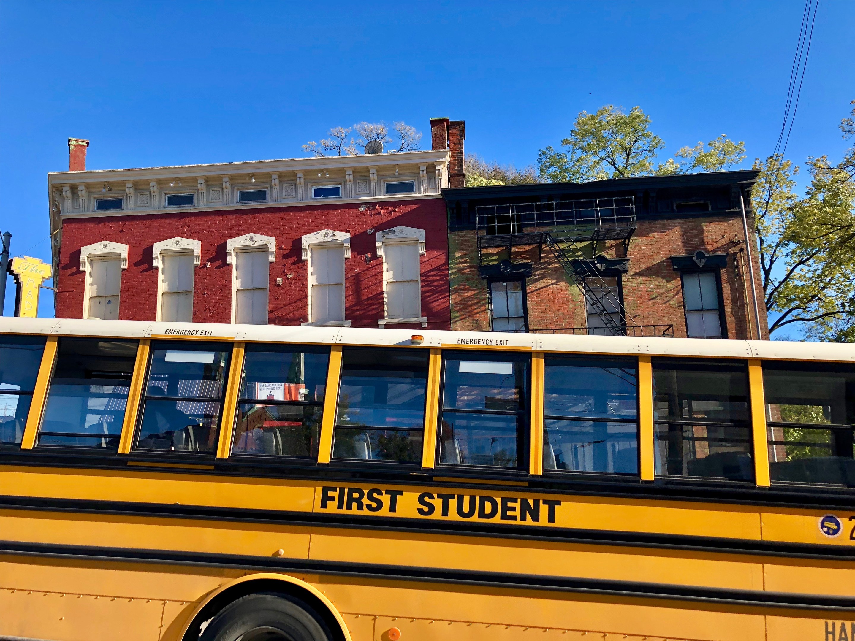 Ein gelber Schulbus der Marke "First Student" vor einem roten Backsteingebäude geparkt, mit ein paar Menschen drinnen und einem klaren blauen Himmel im Hintergrund.