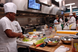 Eine Gruppe von Köchen in einer Küche bei der Zubereitung von Speisen auf einem Schneidbrett, mit einem Koch im Vordergrund in einem weißen Kleid und einer Haube, der ein Messer hält und von verschiedenen Utensilien, Schüsseln, Gemüse, einem Mobiltelefon, Flaschen und anderen Gegenständen umgeben ist, mit einem Fernseher und Deckenlampen im Hintergrund.