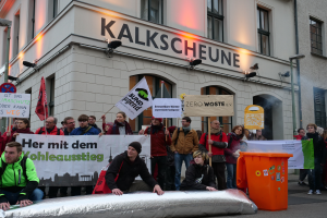 Eine Gruppe von Menschen steht vor einem Gebäude mit Schildern und Plakaten, zwei Personen sitzen im Vordergrund und ein Müllcontainer rechts, während einer Demonstration in Deutschland, mit Gebäuden, Fenstern, Lichtern und Schildern im Hintergrund.
