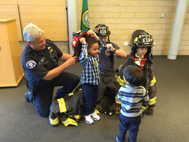 Eine Gruppe von Kindern in Feuerwehruniformen und ein Feuerwehrmann in Uniform stehen um einen Mann in Feuerwehruniform mit einem Helm, einer Tasche auf dem Boden in einer Feuerwache.