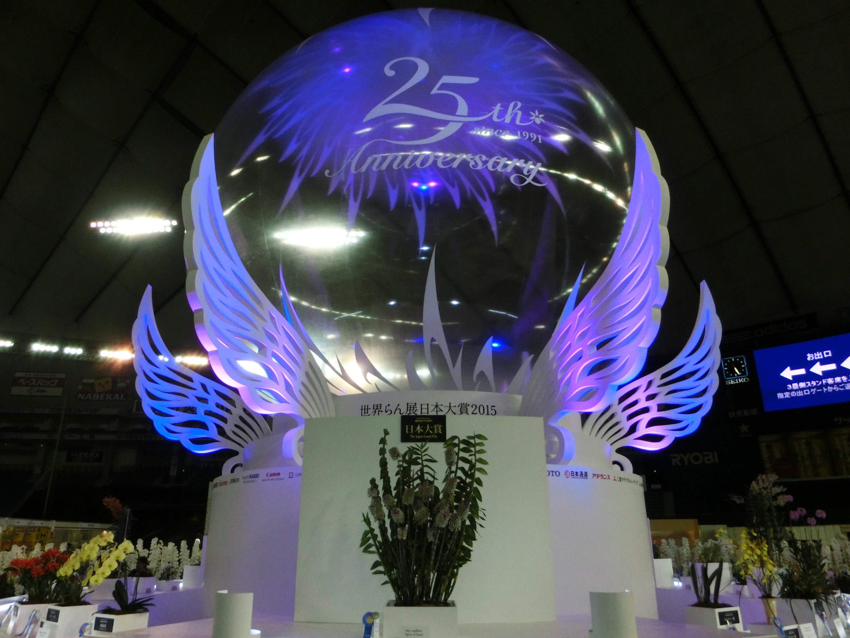 Ein großer Kristallball mit Flügeln oben drauf, umgeben von Blumenbouquets und Namensschildern auf dem Boden, vor einer festlichen Kulisse mit Brettern, Lichtern und einem Bildschirm für ein 25-jähriges Jubiläum.