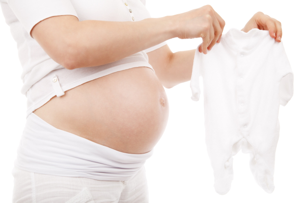 Eine schwangere Frau in einem weißen Kleid hält ein weißes Babyzäpfchen und strahlt glücklich.