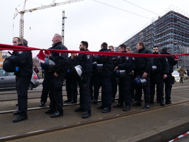 Eine Gruppe von Polizisten in schwarzen Uniformen und Helmen steht in einer Reihe auf einem Bahngleis, mit einem roten Band davor, vor einer Kulisse aus Fahrzeugen, Passanten, Gebäuden und einem klaren Himmel.