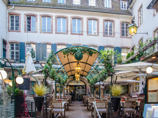 Ein Innenhof mit Tischen und Stühlen, umgeben von Pflanzen, Lichtern, Schildern mit Text, Schirmen und anderen Gegenständen, vor einem Gebäude mit Fenstern unter einem sichtbaren Himmel in Frankfurt, Deutschland.