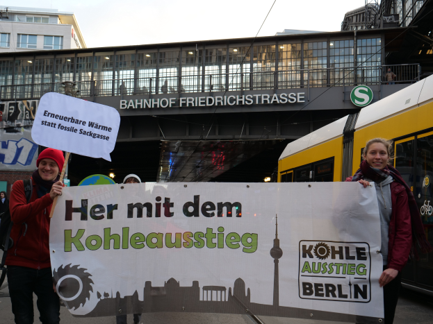 Zwei Personen stehen vor einem Bahnhof und halten ein Schild mit der Aufschrift "Her mit dem Kohleausstieg Berlin"; eine trägt eine rote Jacke und eine rote Mütze, die andere hält eine weiße Tafel, mit mehreren Personen, einem gelben und schwarzen Zug, einer Brücke, Gebäuden, Drähten und dem Himmel im Hintergrund.