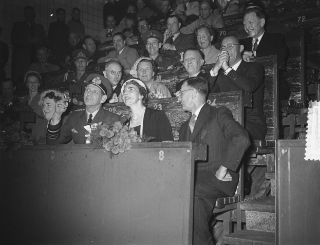 Schwarze und weiße Fotografie einer Menschenmenge in einem Stadion mit Sitzenden und Stehenden, einem Podium mit Blumenstrauß, Stufen auf der rechten Seite und einer Wand im Hintergrund.