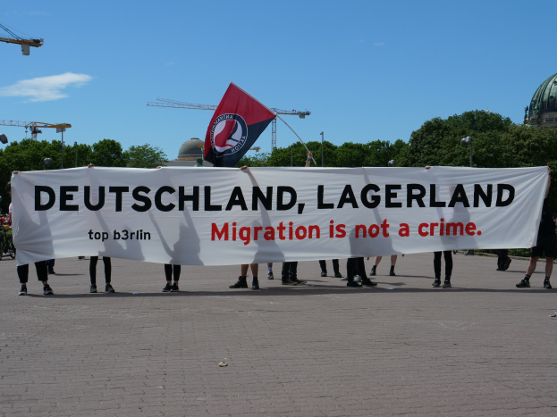 Menschen mit einem Transparent, auf dem 'Deutschland, Lagerland Migration ist kein Verbrechen' steht, und einer Flagge, mit Laternenmasten, Lichtern, Bäumen, Kränen, einem Gebäude und einem bewölktem Himmel im Hintergrund.
