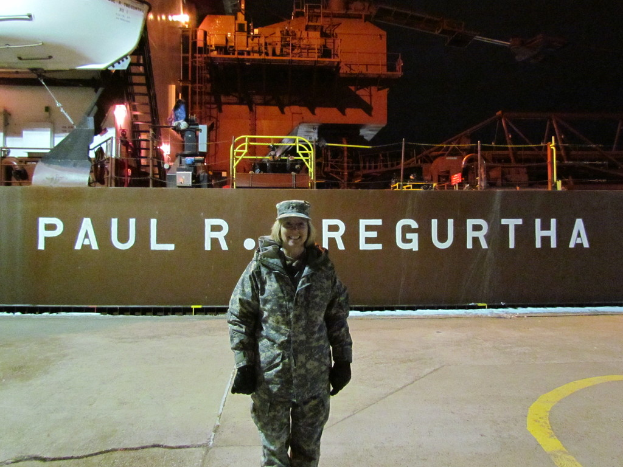 Eine Frau steht lachend mit einer Mütze und Handschuhen vor einem Schiff mit Text im Hintergrund.