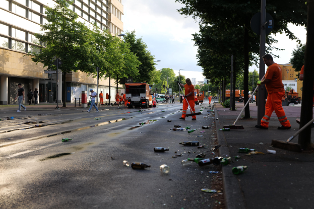 Eine Gruppe von Menschen in orangen Uniformen sammelt Müll von einer Straße, die mit Flaschen und Schutt übersät ist, mit Bäumen, Pfählen und Fahrzeugen im Hintergrund unter einem bewölkten Himmel.