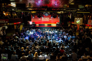 Eine große Menschenmenge sitzt auf Stühlen, die einer Bühne mit Bildschirmen und Plakaten zugewandt sind, beleuchtet von Scheinwerfern und umgeben von Geländern, mit einem Wasserzeichen in der unteren linken Ecke, das eine Atmosphäre der Aufregung und Vorfreude für ein Online-Marketing-Rockstars-Event schafft.
