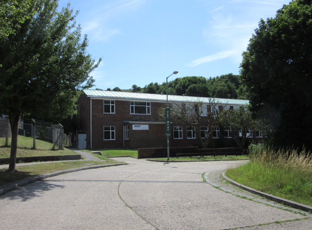 Ein Schulgebäude mit Fenstern, einer Straßenlaterne, einem Schild, einer Baumgruppe, Gras, einem Zaun und einem bewölkten Himmel, das sich in einem Park mit saftigem Grün befindet.