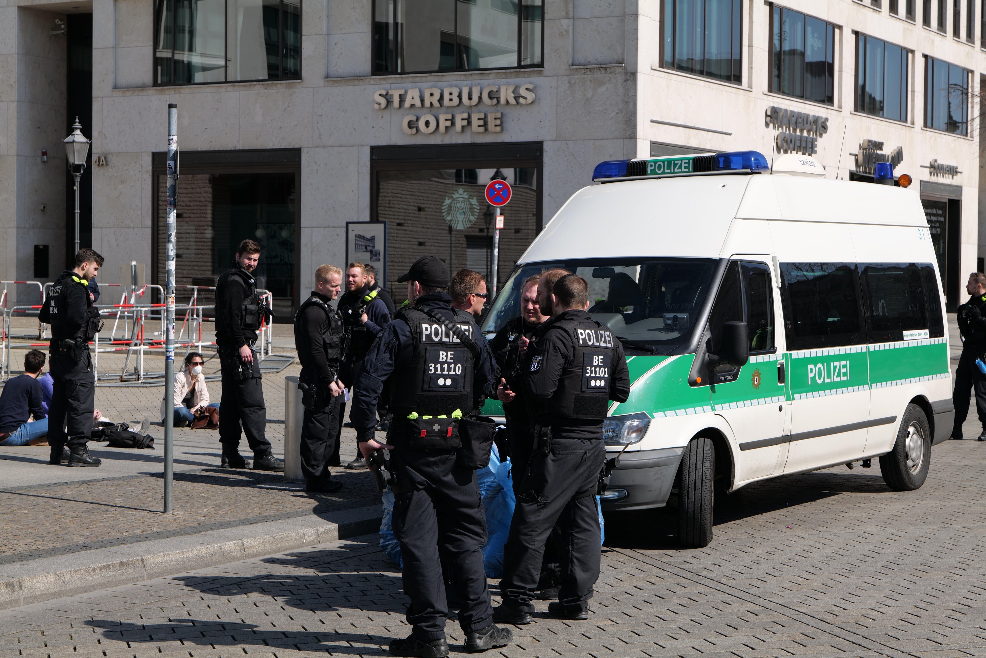 Eine Gruppe von Polizisten steht vor einem Starbucks-Café mit einem Van auf der rechten Seite und einigen Menschen auf der linken Seite, während im Hintergrund ein Gebäude mit Fenstern, ein Schild, ein Laternenpfahl und ein Zaun zu sehen ist.