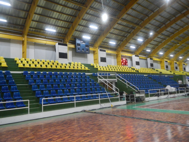 Großer Hallensportplatz mit Holzboden, blauen und gelben Sitzplätzen, Geländern, Treppen mit Geländern, wandmontierten Bildschirmen, Deckenleuchten und wenigen Personen.