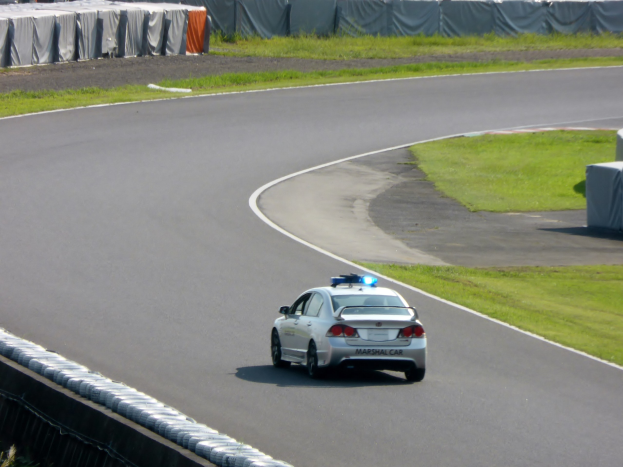 Polizeiauto auf einer Rennstrecke mit Gras auf beiden Seiten und Zuschauern im Hintergrund.