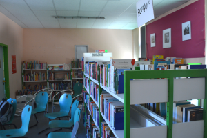 Eine Bibliothek mit Bücherregalen, Stühlen, Taschen auf dem Boden, Rahmen an der Wand, einer Tafel, einer Tür und Deckenleuchten.