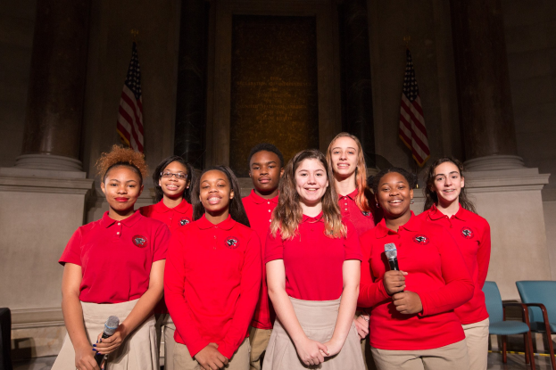 Gruppe junger Frauen in roten Shirts, die strahlend Mikrofone halten und vor Stühlen, Säulen und Fahnen stehen, wahrscheinlich Teil eines nationalen Jugendchors.