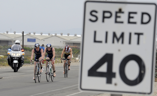 Eine Gruppe von Menschen mit Helmen fährt auf Fahrrädern eine Straße entlang neben einem Geschwindigkeitsbegrenzungsschild, mit Gebäuden, Polen, Pflanzen, Bäumen und einem klaren blauen Himmel im Hintergrund.