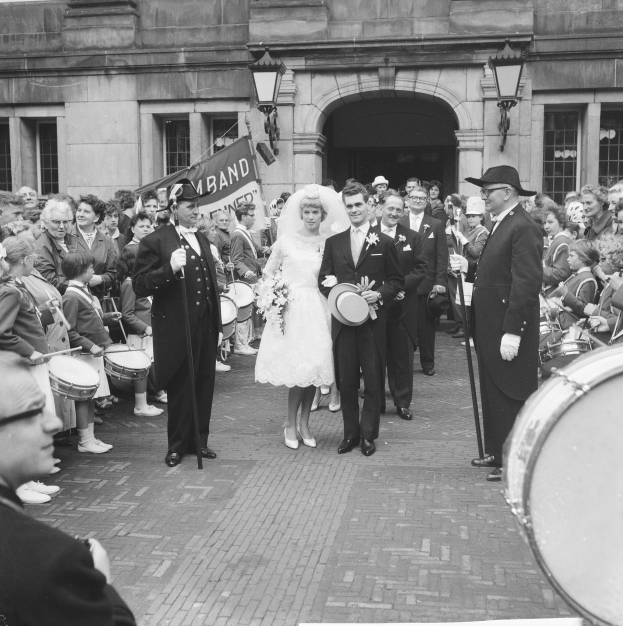 Ein Brautpaar geht eine Straße entlang vor einer Menge, einige halten Musikinstrumente, mit einem Gebäude und einem Banner im Hintergrund, in Schwarz-Weiß.