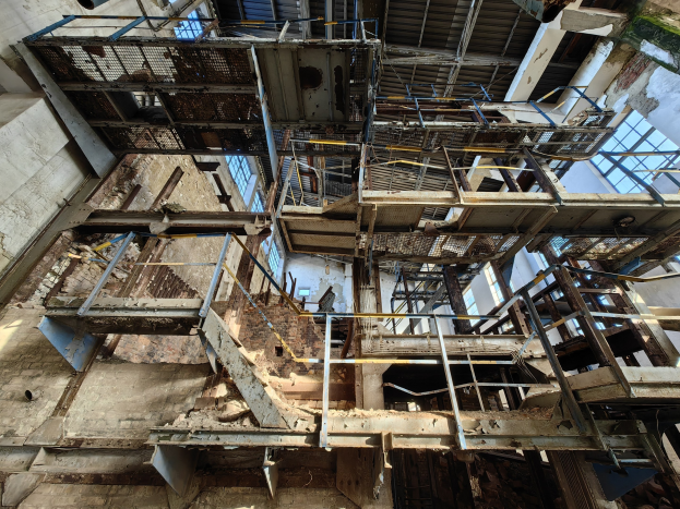 Altes Industriegebäude mit einem zentralen Metallgerüst, verrosteten Wänden, verfallendem Boden und umgeben von Eisenstangen, Treppen und Fenstern.