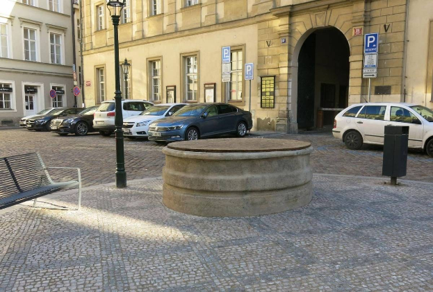 Ein Stadtplatz mit einer Bank in der Mitte, umgeben von Gebäuden, Laternenmasten, Schildern und einem zentralen Springbrunnen.