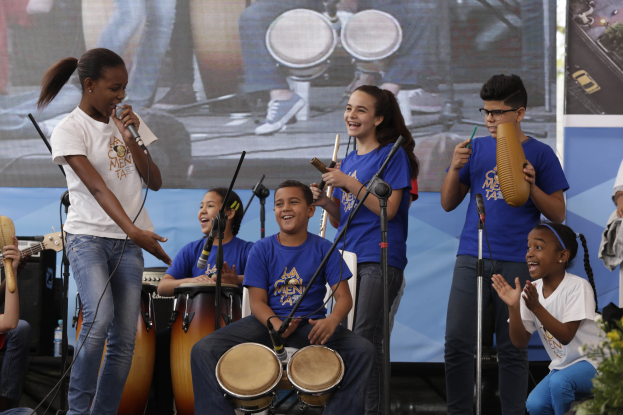 Gruppe von Kindern, die auf einer Bühne Musikinstrumente spielen, mit einer Frau in der Mitte, die ein Mikrofon hält, umgeben von Mikrofonständern, einem Banner im Hintergrund und Pflanzen auf der rechten Seite.