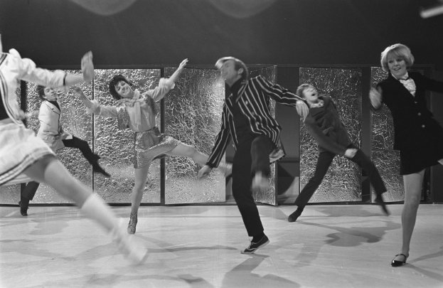 Eine Gruppe von Menschen, die auf einer Bühne tanzen, vor einer Wand, alle in Schwarz-Weiß, mit Schuhen.