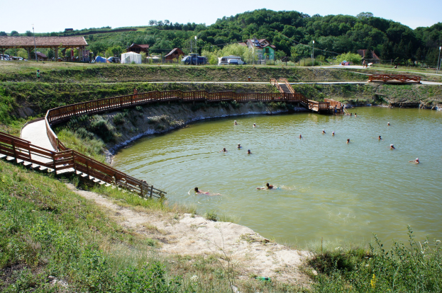 Menschen schwimmen in einem Gewässer mit üppiger Vegetation, einer Brücke mit Treppen, Schuppen, Fahrzeugen, Pfählen und einem klaren blauen Himmel im Hintergrund.
